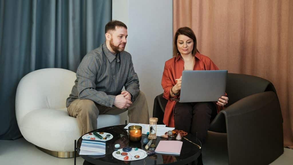 A man and woman sit in chairs around a table while looking at a laptop.
