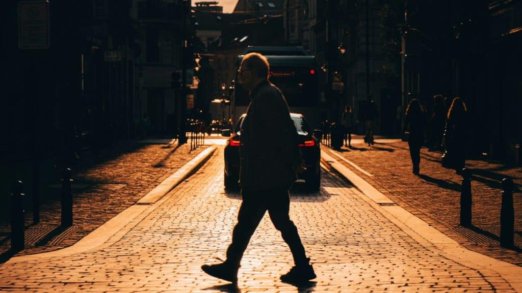 A person walks across a cobblestone street during sunset with buildings and cars in background.