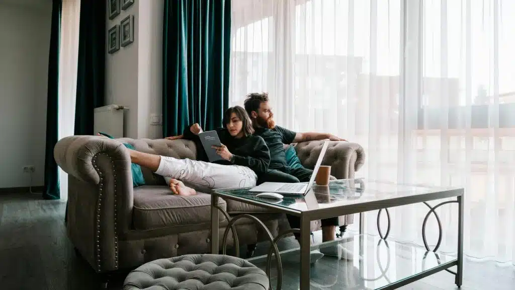 A couple relaxing on a couch with a book and laptop in a bright living room.