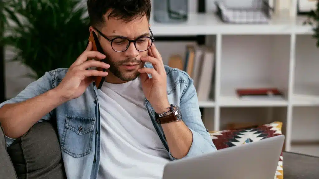 A man wearing glasses talks on the phone while looking at a laptop.