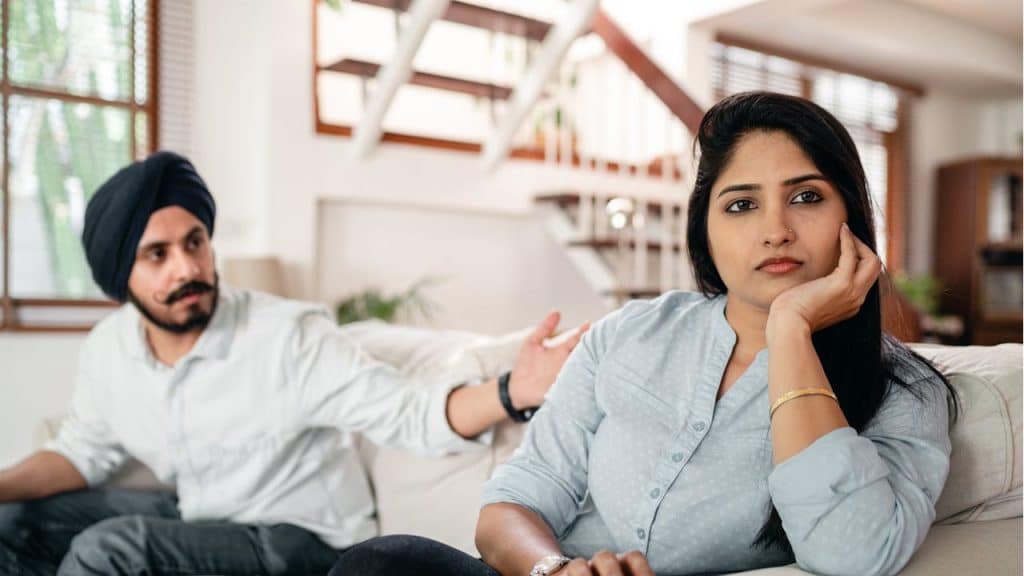 Indian woman looking disgruntled on a sofa while a man in a black turban gestures in the background.