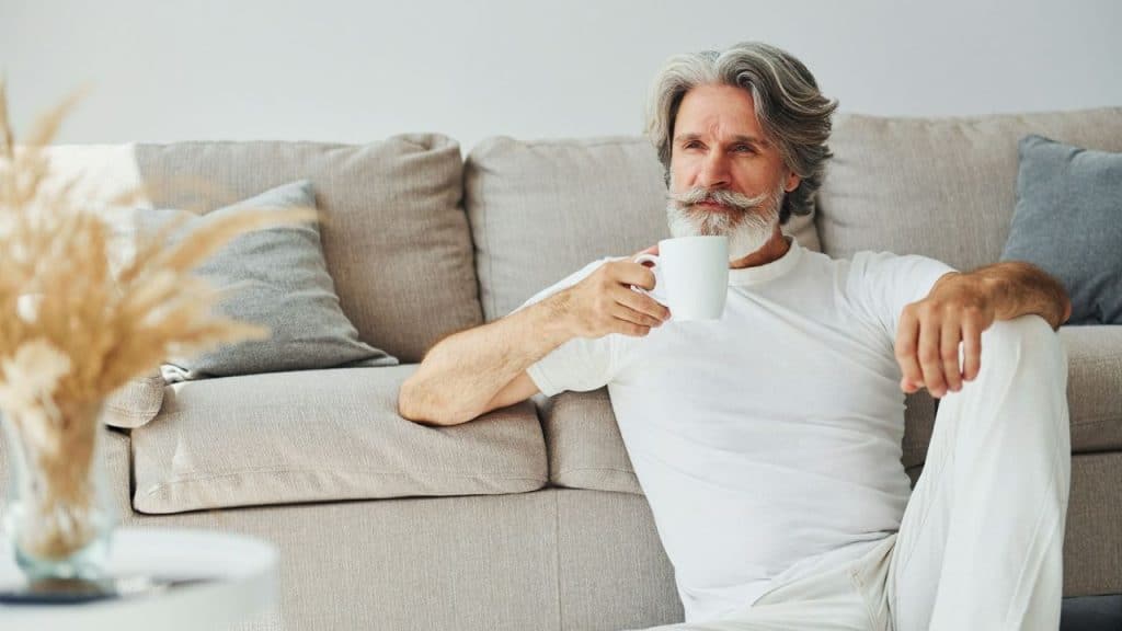 A man with a white beard sitting on the floor holding a white coffee mug.
