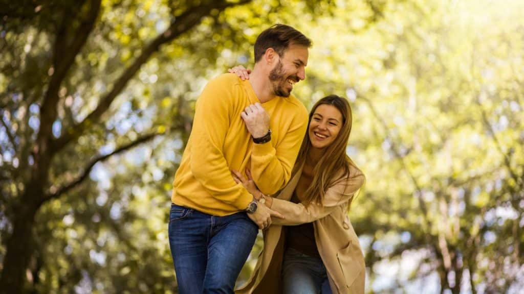 A woman in a tan coat playfully grabs a man in a yellow sweater outdoors.
