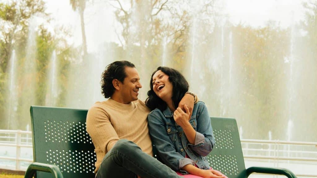 A couple sits on a bench laughing together in front of a fountain.