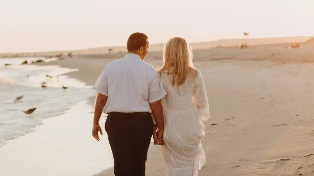 A man and woman walk away from the camera on a sandy beach at sunset.