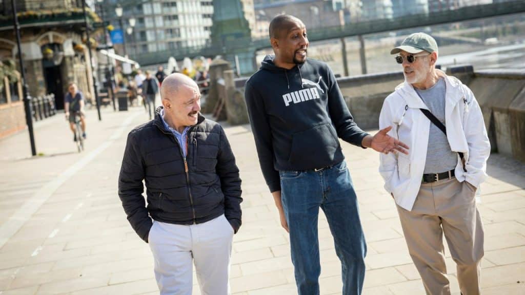 Three men walk together on a sunny paved walkway near a body of water.