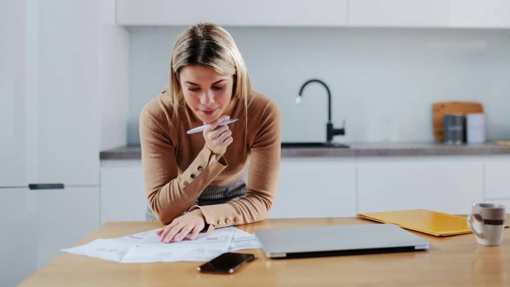 A woman leans over a wooden table looking at papers while holding a silver pen.