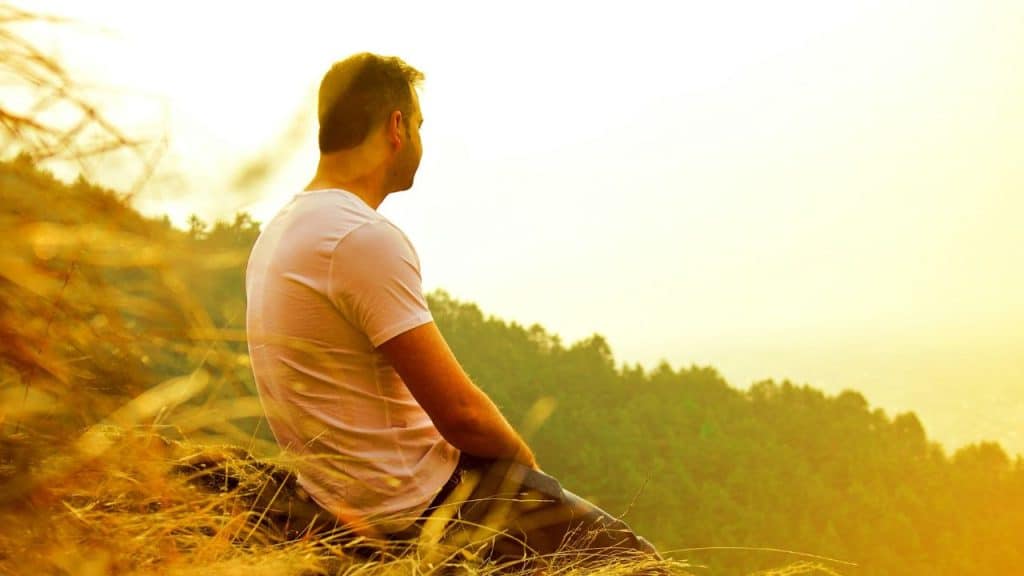 A man in a white shirt sits on a grassy hill looking at a valley.