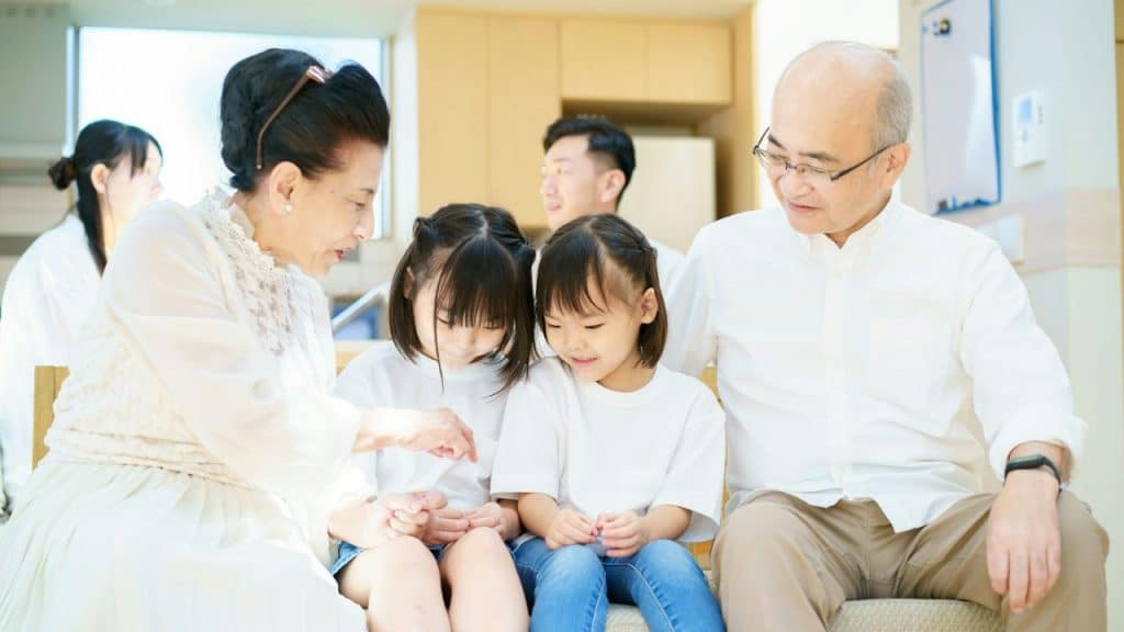 Two older adults and two children sit on a bench together looking at something.