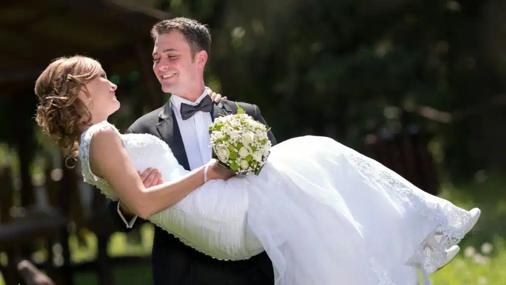 A man in a tuxedo carries a woman in a white lace dress while smiling.