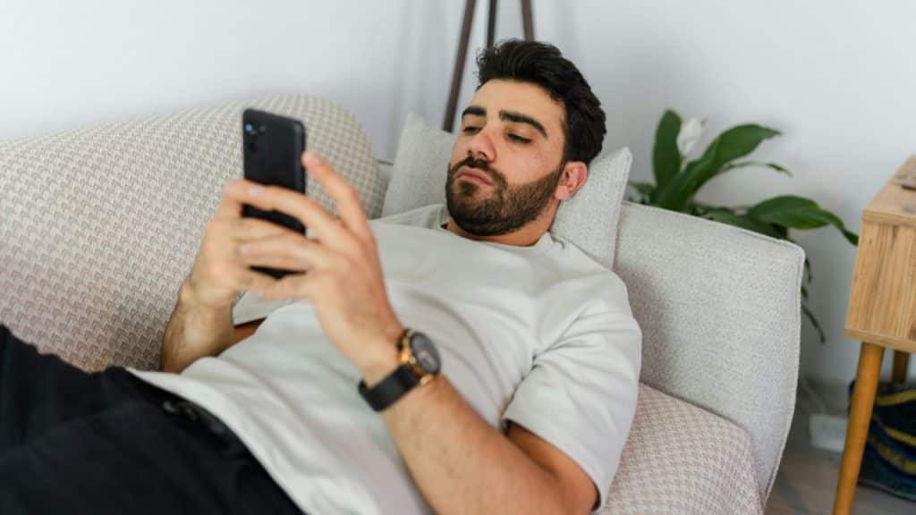 A man with a beard lies on a sofa while looking at a black smartphone.