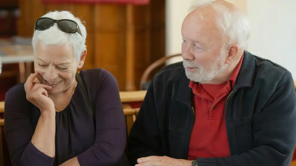 A grey-haired woman with sunglasses on her head laughs beside an older bearded man.
