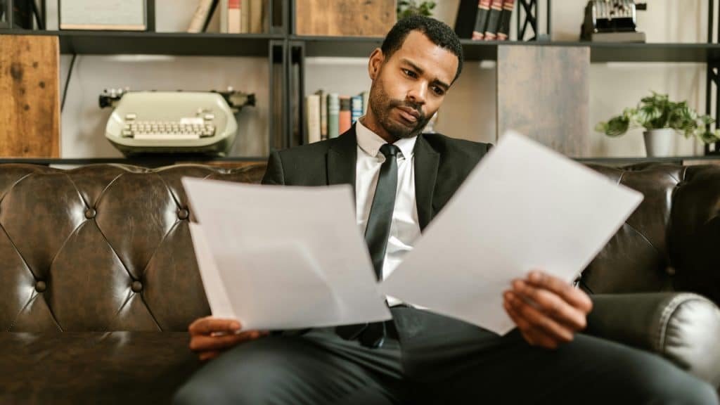 A man in a suit sits on a brown leather sofa, holding two separate documents.