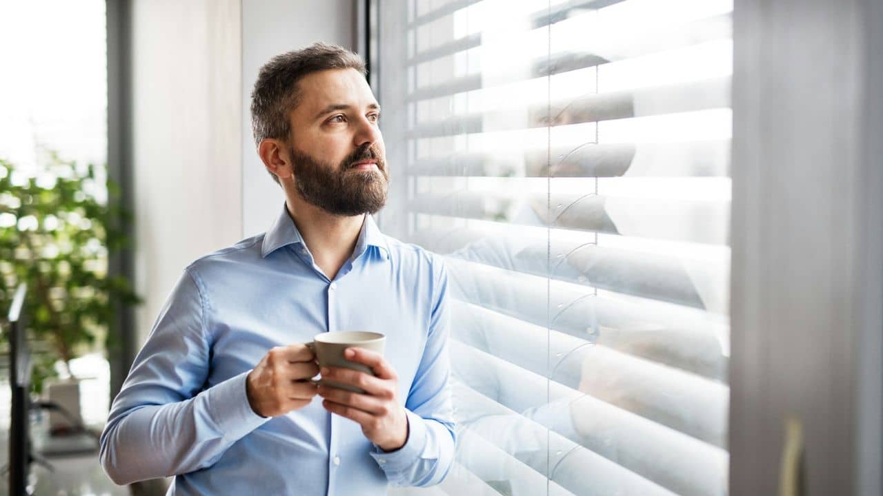 A bearded man in a blue shirt holds a mug while looking out a window's blinds.