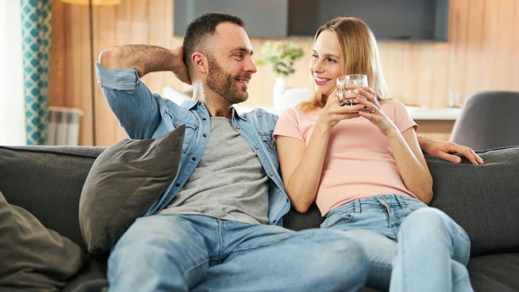 A man and woman sit closely on a dark sofa while the woman holds a glass.