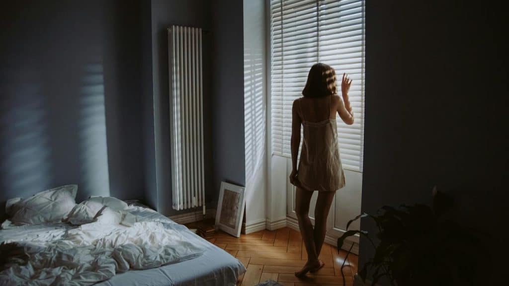 A woman standing by a window looking outside in a dimly lit bedroom.
