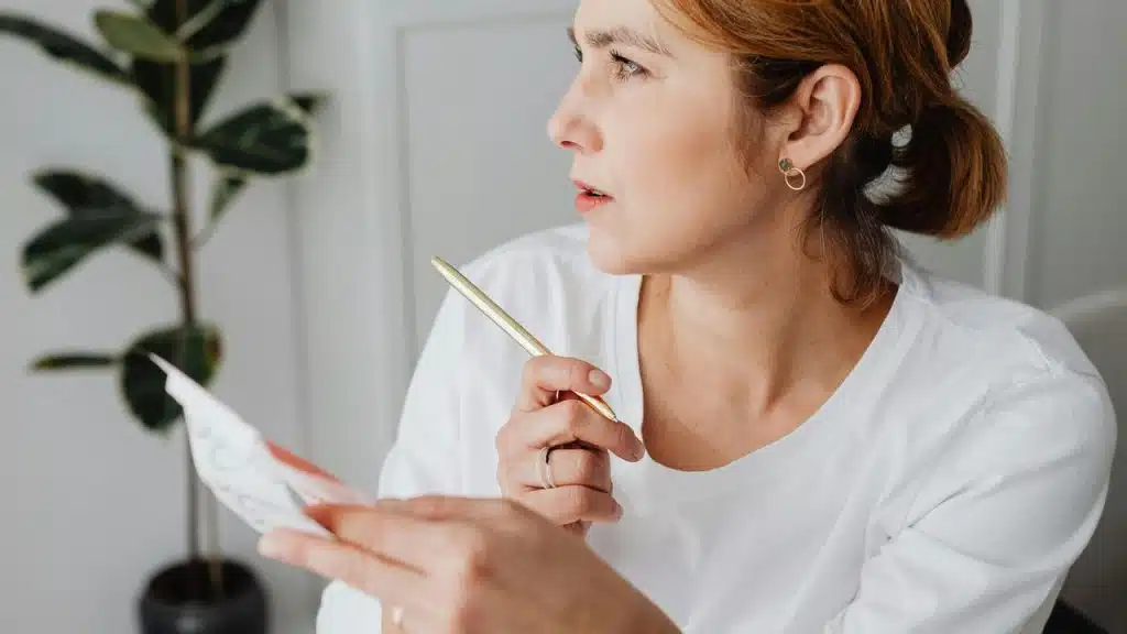 A woman holds a pen and paper while looking to the side.