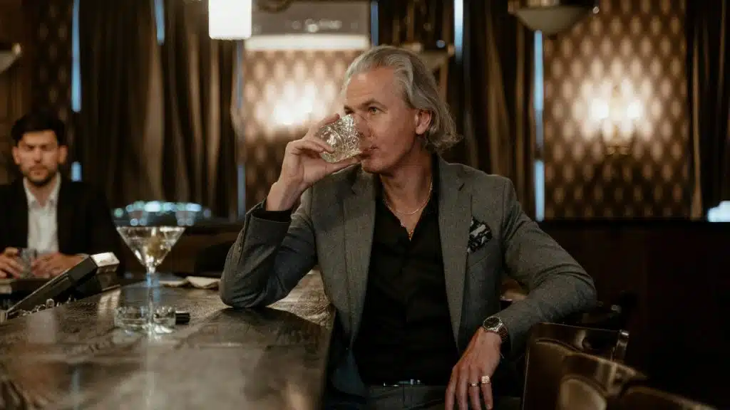 A man in a grey suit sits at a dark bar and drinks from a glass tumbler.
