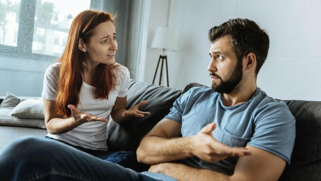 A woman and a man sit on a couch facing each other, both gesturing with their hands and appearing to argue or have an intense discussion in a living room.