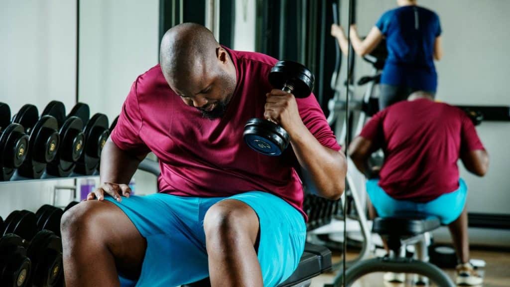 A man in a maroon shirt performs bicep curls while sitting on a gym bench.