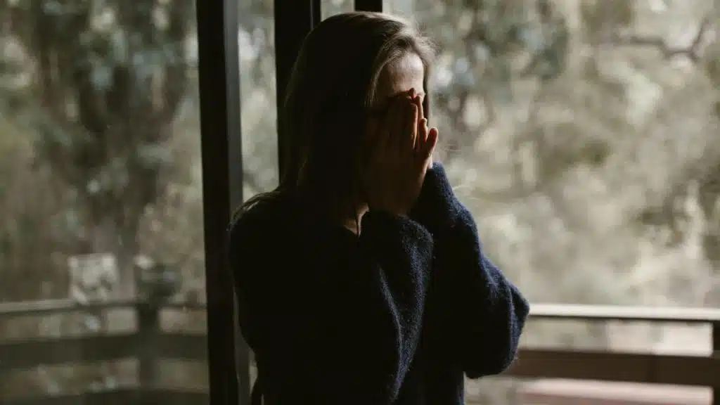 A person standing by a window with their hands covering their face.