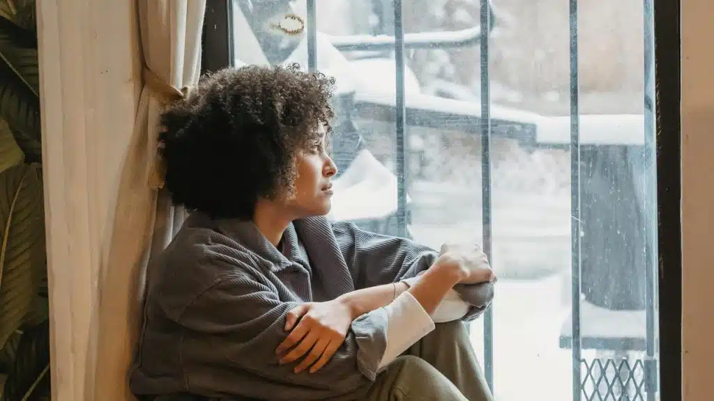 A woman sitting by a window and looking outside with a thoughtful expression.