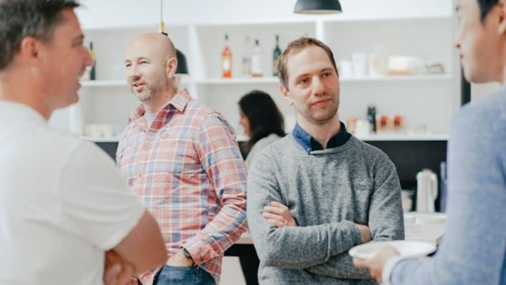 Four men standing in a brightly lit room talking, with one man's arms crossed.