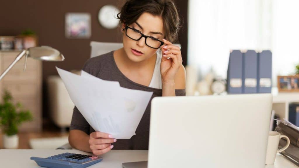 A woman with glasses reviews paper documents while sitting at a desk with a laptop.