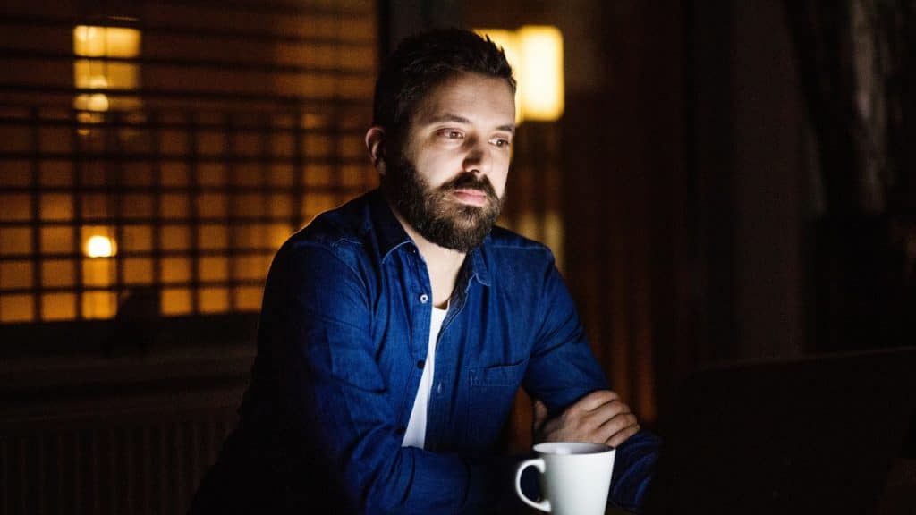 A man with a beard sits in a dark room looking at a glowing laptop screen.