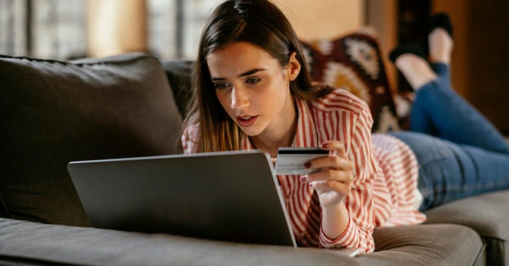 A person lies on a couch using a laptop while holding a credit card.
