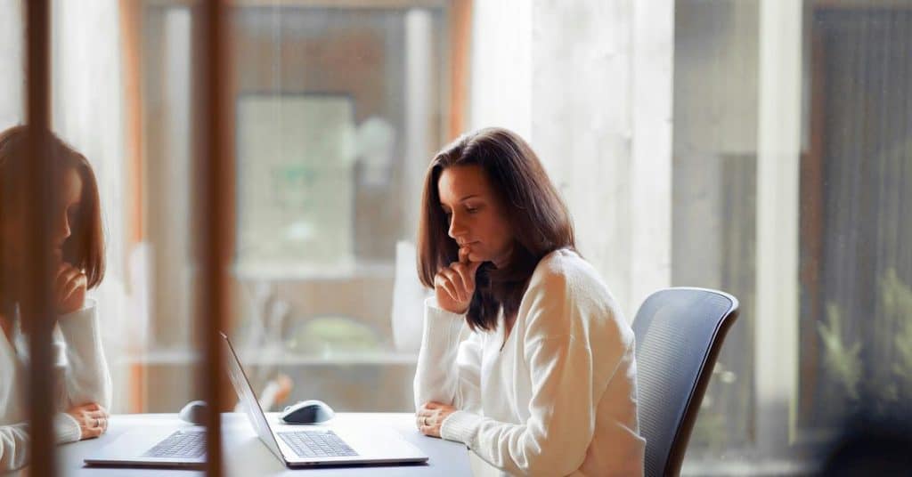 A woman thoughtfully working on a laptop at a desk near a window.