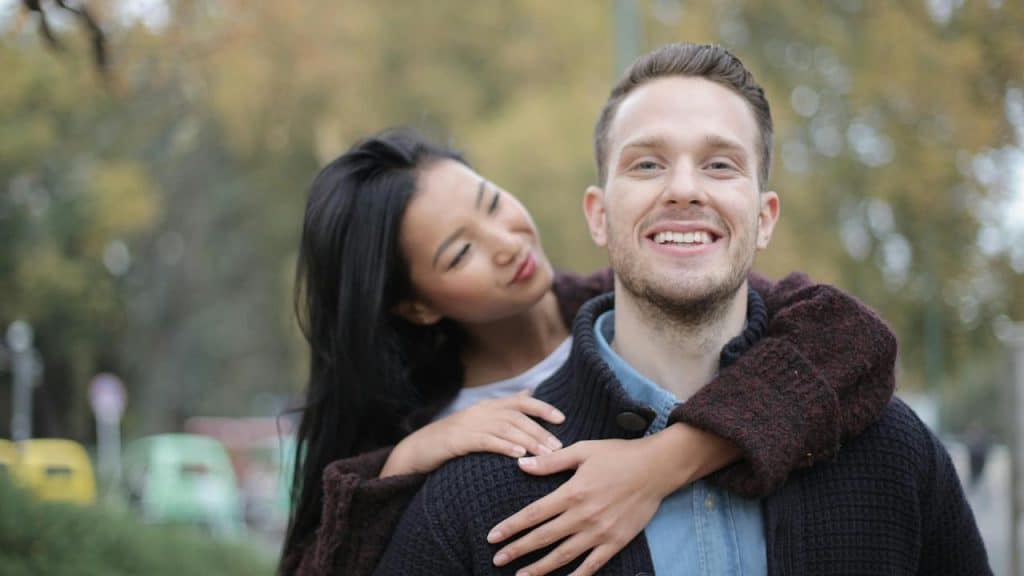 A woman hugging a smiling man from behind outdoors.