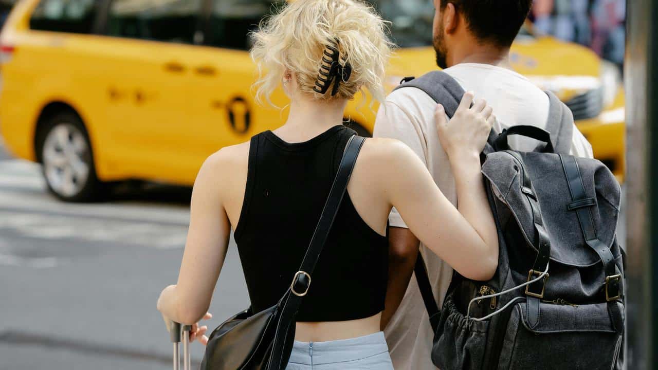 A couple standing on a city street with luggage, waiting beside a yellow taxi.