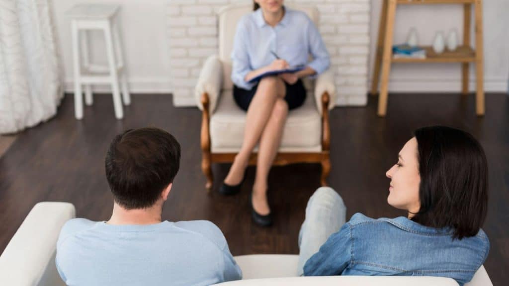 A man and woman sit on a white couch facing a person holding a clipboard.