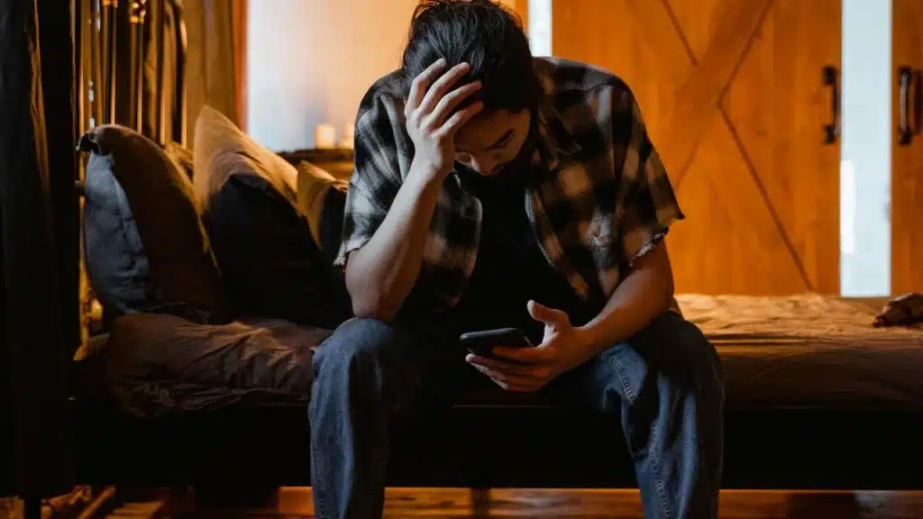 A man sitting on a bed looking at his phone with his head in his hand.