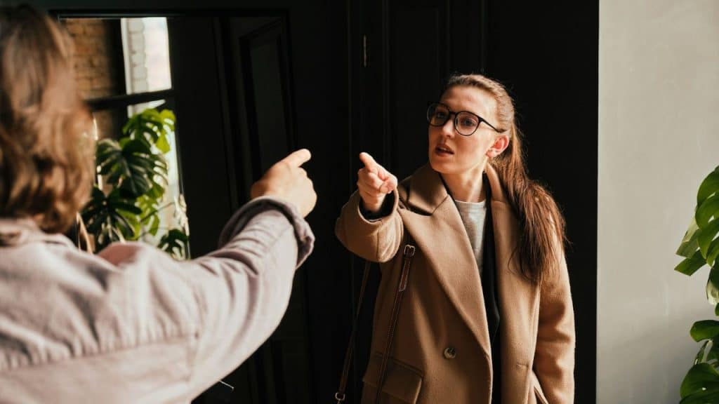 A woman wearing glasses and a tan coat points her finger directly at another person.