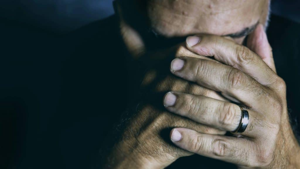 A close-up of a person's hands clasped over their face, featuring a silver wedding band.