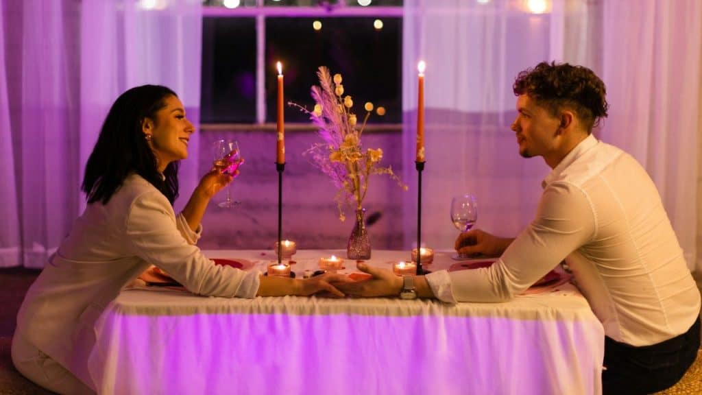 A man and woman sit at a candlelit table holding hands and glasses of wine.