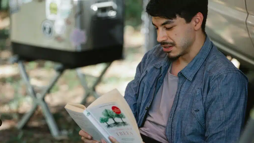 A man in a blue button-down shirt reads a book outdoors near a silver cooler.