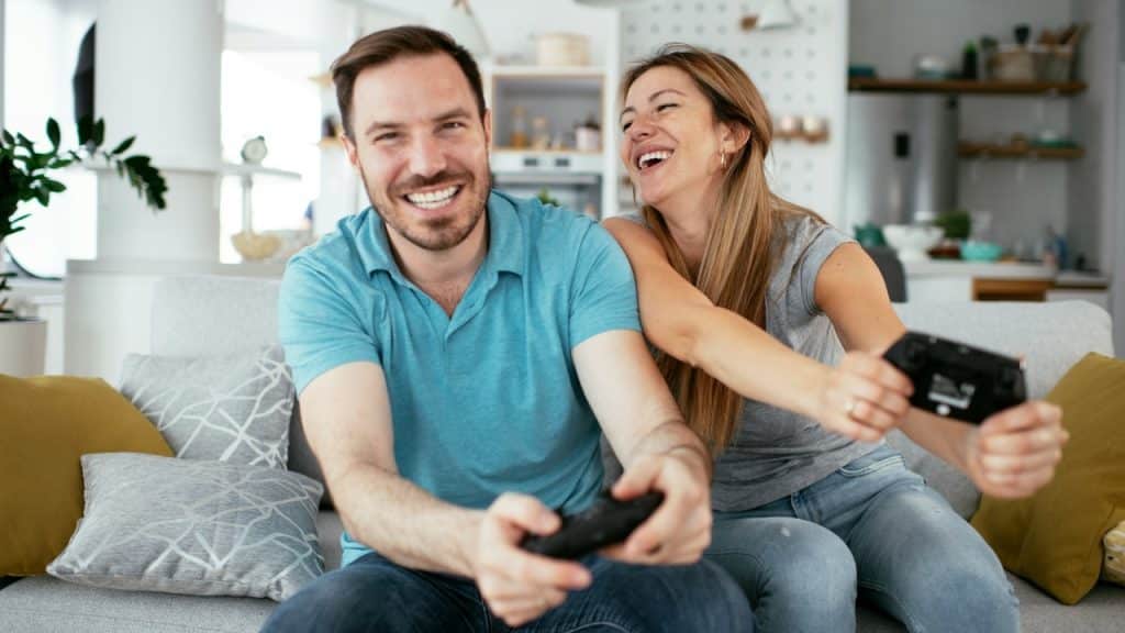 A man and woman sit on a couch laughing while holding black video game controllers.