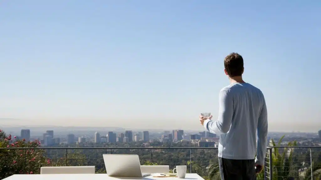 A man standing on a terrace, holding a drink and looking over a city skyline.