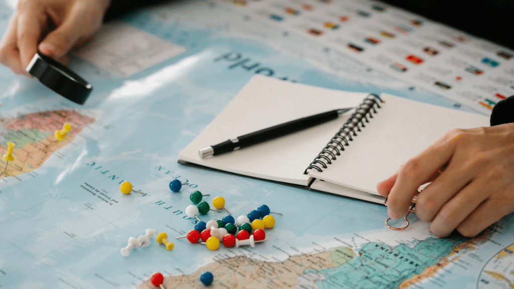 A person’s hands are near a world map, a notebook, a pen, and colorful pushpins.