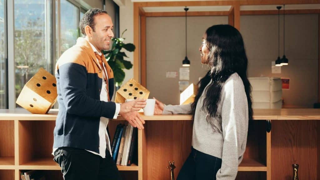 A man and woman stand in an office setting holding mugs and talking to each other.