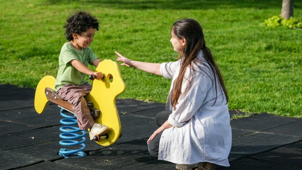 A woman kneels on grass while a young child sits on a yellow spring rider.