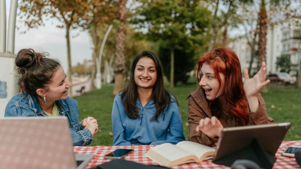 A group of friends laughing together at an outdoor table with laptops and books in a park.
