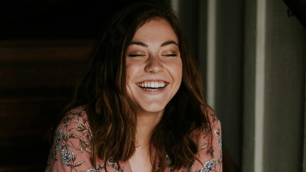A woman with long hair laughing with her eyes closed indoors.