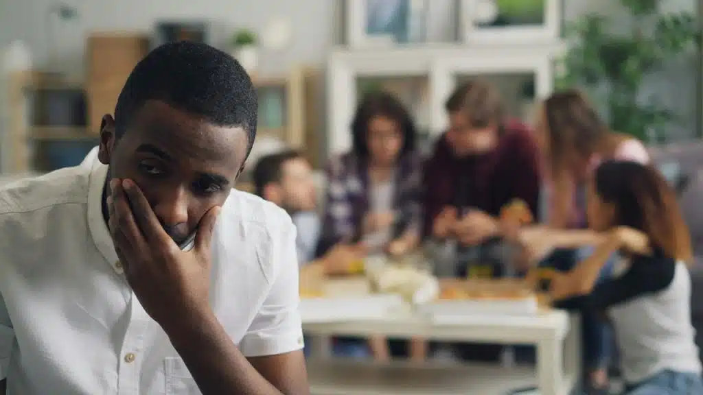 A man sits looking upset with a group of friends socializing in the background.