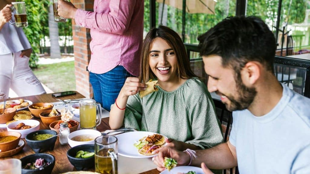 A woman and man enjoy tacos and drinks while laughing at a lively outdoor meal.