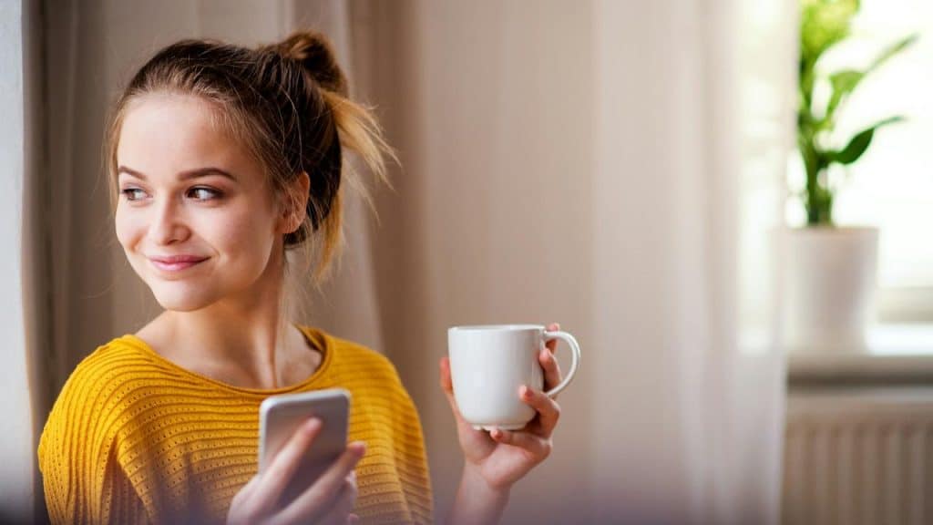 A woman in a yellow sweater holding a phone and a mug while smiling
