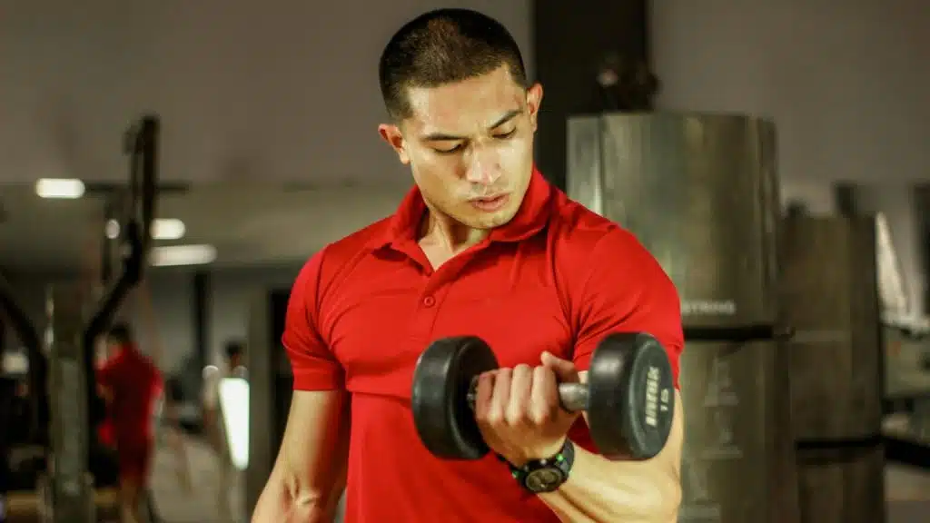 A man in a red polo shirt lifts a dumbbell while looking down in a gym.